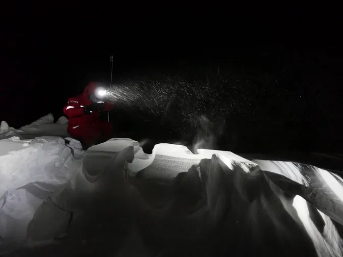 Ein Mann mit einer Stirnlampe sitzt bei Dunkelheit im Schnee,der sich hoch auftäürmt. Schneeverwehungen prägen das Bild im Vordergrund. Es ist grau weiß im Hintergrreund schwarz bis auf den Lichtkegel der Stirnlampe in dem Schneeflocken zu sehen sind. Sie werden von links nach rechts geweht. Der Mann trägt einen dicken roten Schneeanzug.